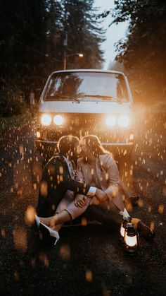 two people sitting on the ground in front of a car with their headlight on