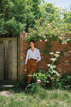 a woman standing in front of a brick wall next to a bush and pink flowers