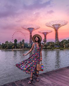 a woman wearing a hat and dress standing on a dock next to water with trees in the background