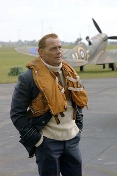 a man in an orange jacket and white sweater standing next to an airplane on the tarmac