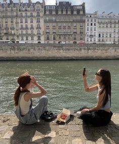 two young women sitting on the edge of a river taking pictures