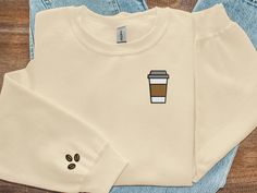 This coffee embroidered crewneck sweatshirt is the perfect blend of style and comfort! Whether you're out and about or cuddled up at home, our crewnecks feature minimalistic designs that can be paired with any outfit. The surprise sleeve design perfectly complements the front, offering a whimsical touch that will make you smile throughout the day! Product Details:  * 50% cotton, 50% polyester * Pre-shrunk * Classic fit * 1x1 athletic rib knit collar with spandex * Air-jet spun yarn with a soft f Coffee Long Sleeve Cotton Top, Coffee Crewneck, Embroidered Coffee Sleeve, Hand Embroidery Coffee Designs, Coffee Shop Sweatshirts, Coffee Crew Neck Top With Funny Print, Coffee Sweater, Embroidery Sweater, Embroidered Crewneck