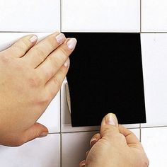 two hands on a white tiled wall with a black square