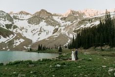 a bride and groom standing in front of a mountain lake with snow on the mountains behind them