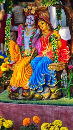 a statue of two people sitting on a swing in front of some flowers and trees