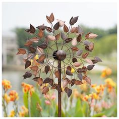 a metal wind spin with leaves on it in front of some yellow and pink flowers