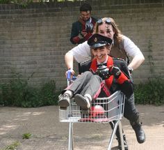 a man in a shopping cart with two other men behind him and one woman sitting on the back