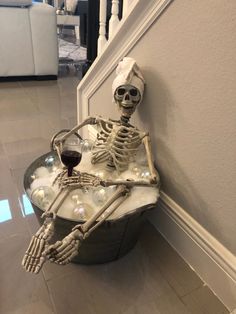 a skeleton sitting on top of a bucket with a glass of wine next to it