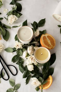 an arrangement of flowers, oranges and scissors on a white tablecloth with candles