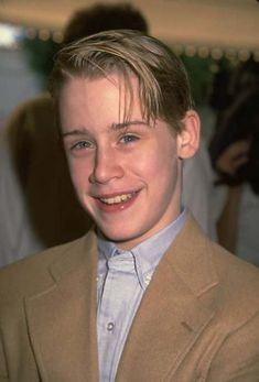 a young man wearing a tan jacket and blue shirt smiles at the camera while standing in front of a crowd