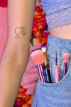 a woman's arm with several makeup brushes in her pocket, and the other half of her arm has a tattoo on it