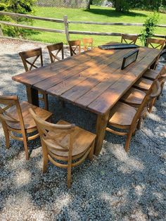 a wooden table with six chairs around it in gravel area next to fence and trees