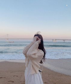 a woman standing on top of a sandy beach next to the ocean with a bridge in the background