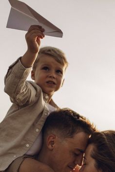 a woman holding a little boy on her shoulders while he holds a paper airplane in the air