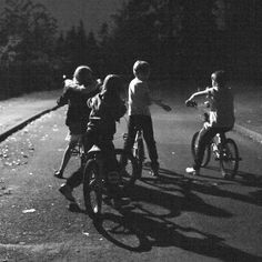 black and white photograph of children riding bikes in the street at night with headlights on