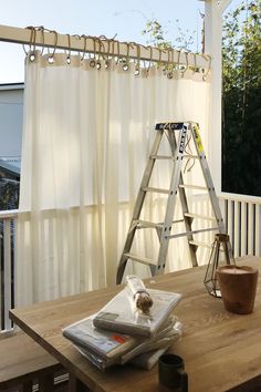 a wooden table topped with books and a ladder next to a window covered in white curtains