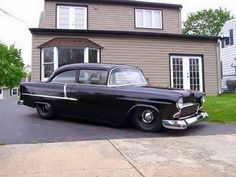 an old black car is parked in front of a house with a brown siding and two windows