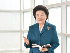 an older woman is reading a book and smiling at the camera while standing in front of large windows