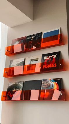 three orange shelves with cd's on them in front of a white wall and window