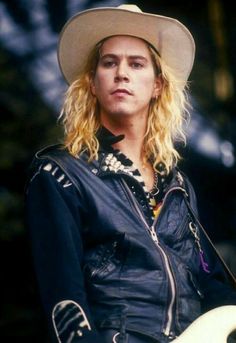 a man with long blonde hair wearing a hat and leather jacket, looking at the camera