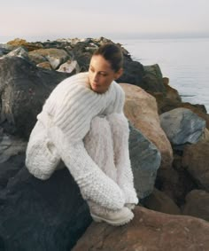 a woman is sitting on some rocks by the water wearing a white sweater and pants