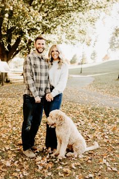 a man and woman holding hands while standing next to a dog in the fall leaves