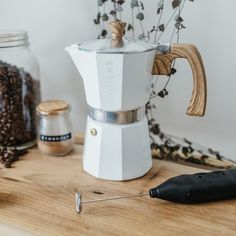 a white coffee maker sitting on top of a wooden table next to beans and other items