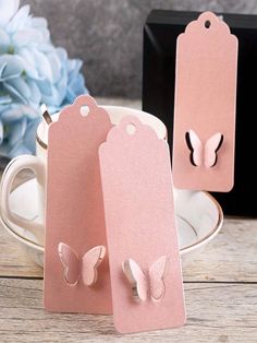 two pink tags with butterflies on them sitting next to a coffee cup and saucer