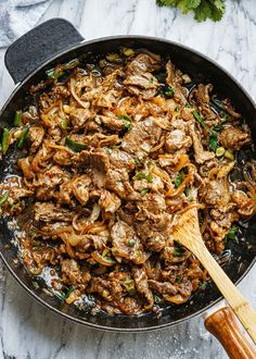 a skillet filled with meat and vegetables on top of a marble counter next to a wooden spatula