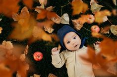 a baby is laying on the ground surrounded by autumn leaves and an apple, looking up at the camera