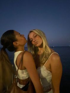 two beautiful young women standing next to each other near the ocean at night with boats in the background