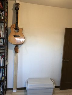 a guitar hanging on the wall next to a trash can and bookshelf in a room