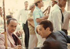 two men sitting at a table playing cards in front of other people on the street