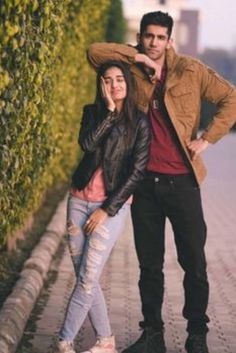 a man and woman standing next to each other on a sidewalk with hedges behind them