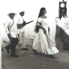 a woman in a white dress and two men are standing on the sidewalk with guitars