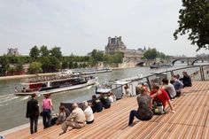 people are sitting on the edge of a wooden pier as boats pass by in the distance