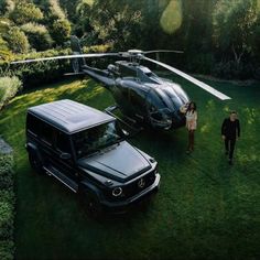 two people standing in front of a black suv next to a helicopter on the grass