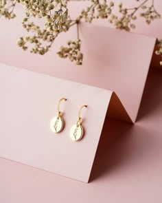 two small gold earrings sitting on top of a pink table next to some white flowers