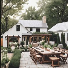 an outdoor dining area with wooden tables and chairs in front of a white farmhouse style house