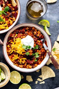 two bowls filled with chili, beans and cheese next to tortilla chips on a table