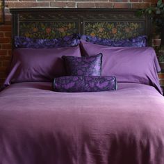 a bed with purple sheets and pillows in front of a brick wall next to a potted plant