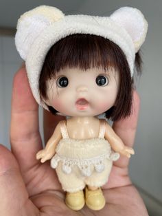 a hand holding a small doll in it's right hand and wearing a white hat