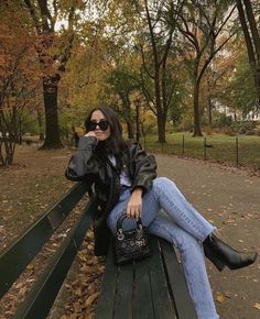 a woman is sitting on a park bench talking on her cell phone and holding a purse