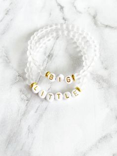 two white and gold bracelets with the words big little on them sitting on a marble surface