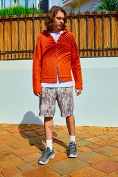 a man standing in front of a fence wearing an orange sweater and shorts with white socks