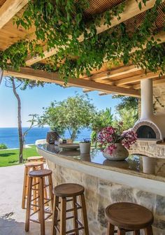 an outdoor kitchen with stone counter tops and wooden stools under a pergolated roof