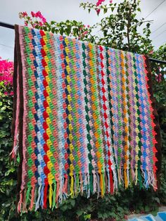 a multicolored crocheted blanket hanging on a clothes line with flowers in the background