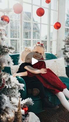 two children hugging each other on a couch in front of a christmas tree with red balls