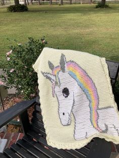 a crocheted unicorn blanket sitting on top of a wooden bench next to a bush