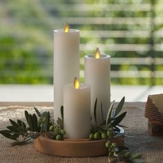 three white candles sitting on top of a wooden stand next to green leaves and berries